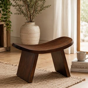 Op de afbeelding: Een donkerbruine houten kruk met een gebogen zitting en schuine poten. De kruk staat op een geweven vloerkleed. Een beige mok en boeken staan in de buurt, wat wijst op een gezellig, minimalistisch interieur.