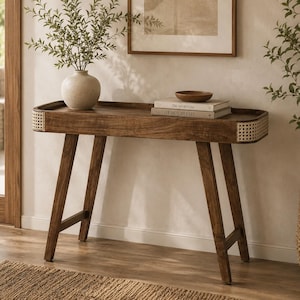 May include: A rectangular wooden console table with a dark brown finish, featuring rounded ends with light-colored woven cane detailing. The table has four angled legs and a flat top surface, currently holding a beige ceramic vase and two books.