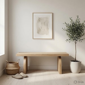 May include: A light-toned wooden bench with curved legs, positioned below a framed abstract line art print. To the left sits a woven seagrass basket and a pair of beige slippers, while a potted olive tree stands to the right on the floor.
