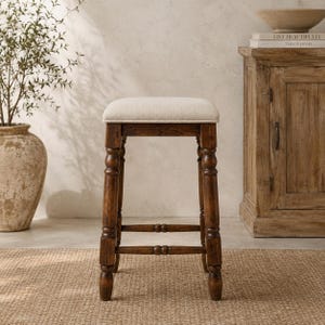 Könnte beinhalten: Ein quadratischer Hocker mit einem dunkelbraunen Holzrahmen, gedrechselten Beinen und horizontalen Stützstreben. Die Sitzfläche ist mit einem hellbeigen Stoff mit dezenter Webstruktur bezogen. Der Hocker steht auf vier Beinen mit abgerundeten Füßen.