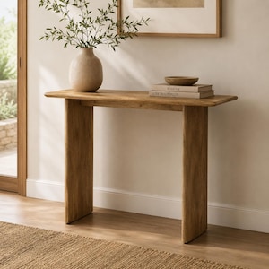 Peut inclure: Une console en bois brun clair avec un plateau rectangulaire et deux pieds verticaux larges et plats. Sur la table se trouve un vase en céramique beige avec des branches feuillues vertes et deux livres empilés intitulés Kinfolk et Live Beautifully.