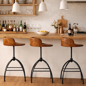 May include: Three adjustable bar stools with wooden seats and black metal bases are arranged in front of a wooden countertop. The stools have a vintage industrial design. The kitchen setting includes white pendant lights and a white tile backsplash.