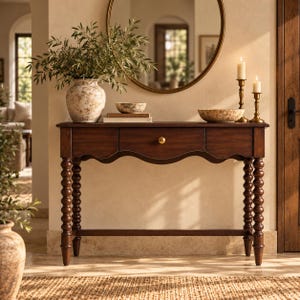 May include: A dark wood console table featuring a central drawer with a small gold knob, supported by four turned spindle legs. The table surface holds a large textured vase with greenery, a small bowl, two books, and two brass candle holders.