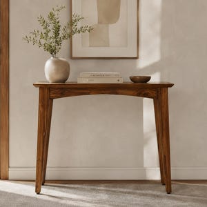 May include: A wooden console table with four tapered legs and an arched apron. On the surface sits a round ceramic vase containing green foliage, two stacked books with the word KIN KIN FOLK visible on the spine, and a small dark wooden bowl.