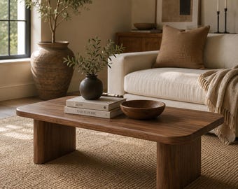 May include: A low-profile rectangular coffee table made of dark brown wood with rounded corners and two thick slab legs. On the table sits a small black vase with green foliage, two stacked books, and a round wooden bowl with a smooth finish.