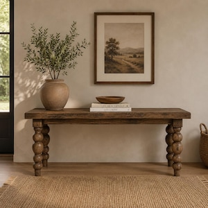 May include: A rectangular wooden bench with four turned bobbin-style legs. On the bench sits a large, round, tan ceramic vase containing green leafy branches, a small wooden bowl, and two stacked books titled Live Beautifully and This Is Home.