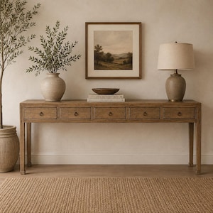 Op de afbeelding: Een lange houten sidetable met vijf lades, elk voorzien van een kleine ronde metalen greep. Op de tafel staan een donkere houten schaal, twee op elkaar gestapelde boeken met de tekst Live Beautifully en een tafellamp met een beige kap.