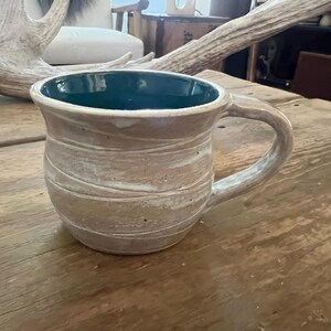 May include: Handmade ceramic mug with a beige exterior and a teal interior. The mug features a textured, wrapped design and a curved handle. It sits on a wooden surface, showcasing its rustic charm. A unique piece for enjoying beverages.