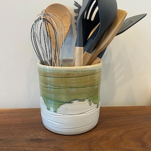 May include: A ceramic utensil holder with green and white glaze, containing kitchen tools. The tools include a whisk, wooden spoons, spatulas, and a ladle. The holder is cylindrical and rests on a wooden surface.