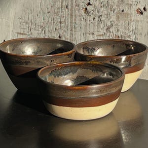 Könnte beinhalten: Drei Keramikschalen mit glänzendem Finish. Die Schalen haben einen dunkelbraunen Innenraum, ein braunes Band und einen cremefarbenen Boden. Die Schalen sind in einer Gruppe angeordnet, wobei die kleinste Schale vorne steht. Der Hintergrund ist eine verwitterte Holzoberfläche.