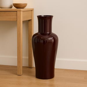 May include: A tall, glossy burgundy vase with a wide base and flared top. The vase is positioned on a light wood floor, near a wooden side table with a lamp and a wooden bowl.