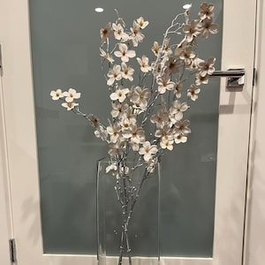 May include: A tall, clear glass vase holding a floral arrangement. The arrangement features branches with small, delicate white flowers. The branches are silver, and the vase is rectangular. The background includes a white door and a gray wall.