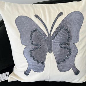 May include: Cream-colored square pillow with a large, gray butterfly design. The butterfly has textured wings and is embellished with dark gray beads. The pillow is on a black surface.