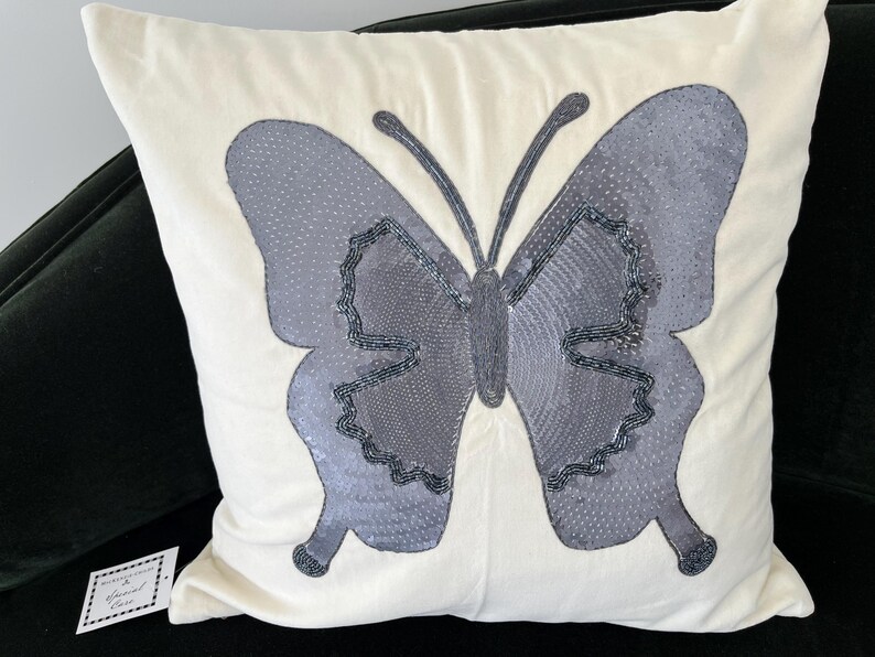 May include: Cream-colored square pillow with a large, gray butterfly design. The butterfly has textured wings and is embellished with dark gray beads. The pillow is on a black surface.