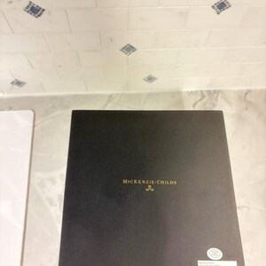 May include: A square black box with the gold text MacKenzie-Childs printed in the center. A small white circular sticker is located in the bottom right corner, and a rectangular white label with text is positioned below the circular sticker.