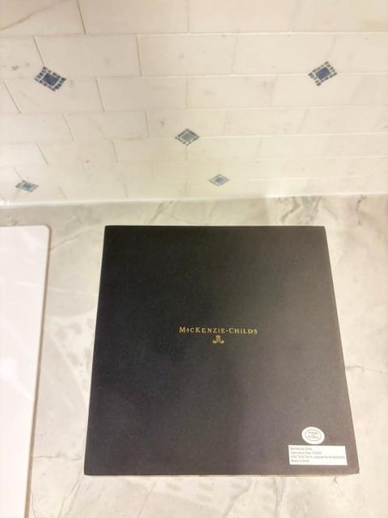 May include: A square black box with the gold text MacKenzie-Childs printed in the center. A small white circular sticker is located in the bottom right corner, and a rectangular white label with text is positioned below the circular sticker.