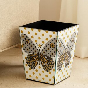 May include: A square wastebasket with a white background featuring a repeating pattern of gold polka dots. The front panel displays a large butterfly graphic with grey, black, and gold wings accented by small, clear, circular embellishments.