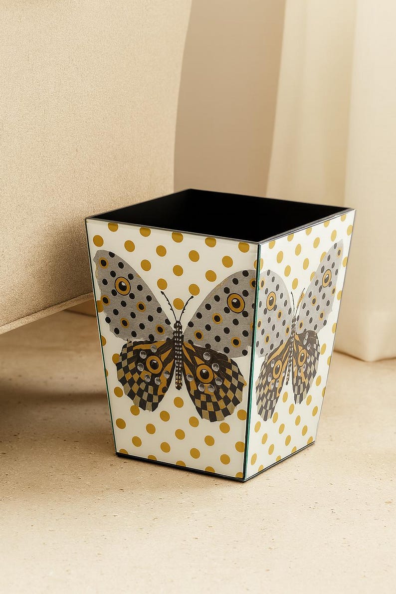 May include: A square wastebasket with a white background featuring a repeating pattern of gold polka dots. The front panel displays a large butterfly graphic with grey, black, and gold wings accented by small, clear, circular embellishments.