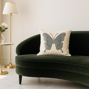 May include: A decorative pillow with a gray butterfly design on a cream-colored background rests on a dark green velvet sofa. A gold lamp and side table with flowers are in the background, creating a cozy living room scene.