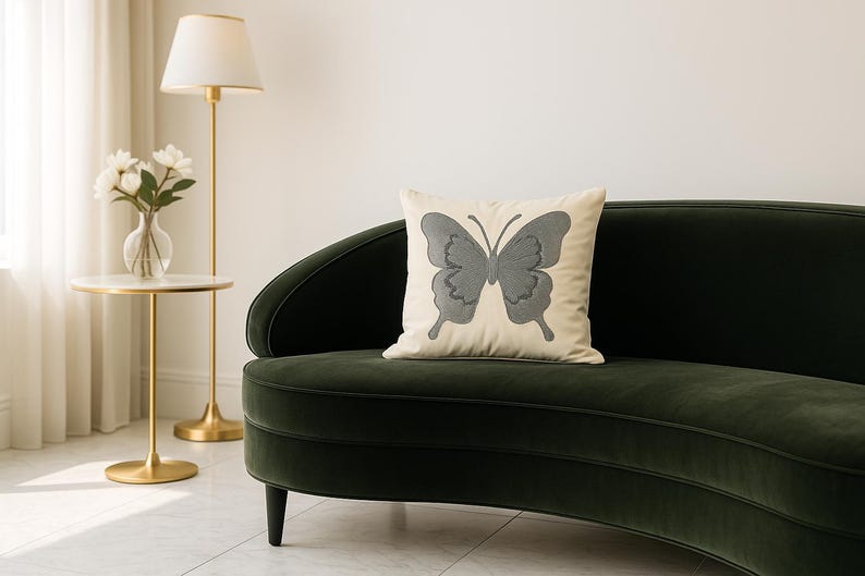 May include: A decorative pillow with a gray butterfly design on a cream-colored background rests on a dark green velvet sofa. A gold lamp and side table with flowers are in the background, creating a cozy living room scene.