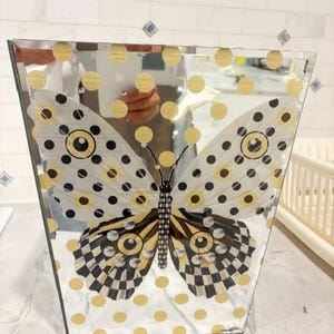May include: A trapezoidal mirrored vase featuring a large butterfly graphic with black, white, and yellow patterns. The surface is decorated with a repeating grid of gold polka dots. The mirrored glass reflects the surrounding environment clearly.