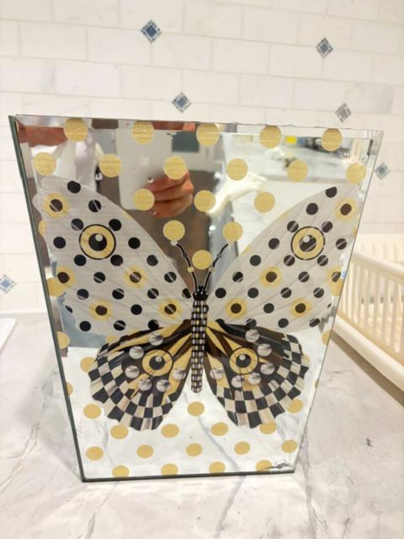 May include: A trapezoidal mirrored vase featuring a large butterfly graphic with black, white, and yellow patterns. The surface is decorated with a repeating grid of gold polka dots. The mirrored glass reflects the surrounding environment clearly.