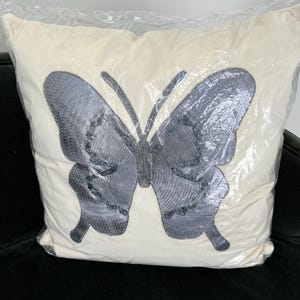 May include: A decorative pillow with a large butterfly design. The butterfly is gray with beaded accents and a textured pattern, set against a cream-colored background. The pillow is encased in clear plastic.