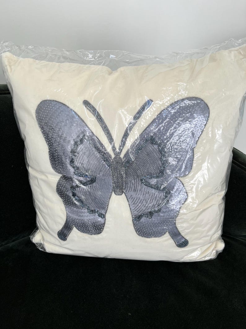May include: A decorative pillow with a large butterfly design. The butterfly is gray with beaded accents and a textured pattern, set against a cream-colored background. The pillow is encased in clear plastic.