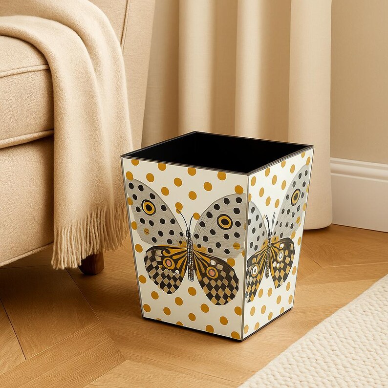 May include: A square wastebasket featuring a white background with a repeating pattern of gold polka dots. The front panel displays a large butterfly illustration with grey, black, and gold wings, featuring circular eye-like markings and checkered patterns.