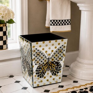 May include: A square wastebasket with a mirrored surface featuring a gold polka dot pattern and a large central butterfly graphic. The butterfly has black, white, and yellow wings with intricate patterns. The interior of the wastebasket is black.