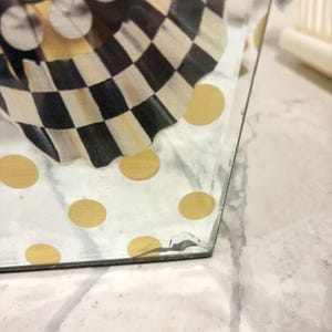 May include: A clear glass container featuring a decorative pattern of gold polka dots on the exterior and a black and white checkered design on the interior. The glass structure shows a visible crack near the bottom corner edge of the object.