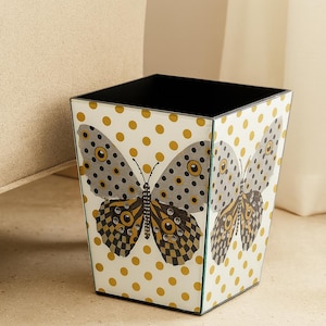 May include: A square wastebasket with a white background featuring a repeating pattern of gold polka dots. The front panel displays a large butterfly graphic with grey, black, and gold wings accented by small, clear, circular embellishments.