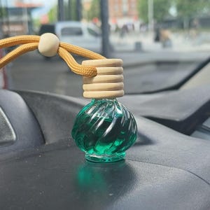 May include: A small, clear glass car air freshener with a swirling green liquid inside. The bottle has a wooden cap and a tan cord for hanging. The air freshener is sitting on a black surface.