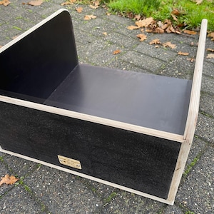 May include: A wooden box with a black interior and exterior, featuring a curved back and a handle. The box has a rectangular shape with a light-coloured wood trim. The box has a small rectangular label on the side.