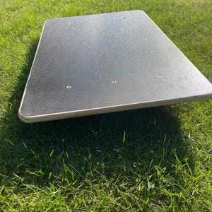 Könnte beinhalten: Ein rechteckiges Balance Board mit einer schwarzen strukturierten Oberfläche und abgerundeten Kanten. Das Board ist aus Holz gefertigt und hat drei kleine Löcher. Das Board liegt auf einer grünen, grasbewachsenen Oberfläche.
