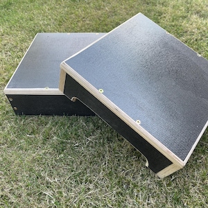 May include: Two black wooden step platforms with a textured surface and light-coloured wood trim. The platforms are rectangular and are placed on a grassy surface. The platforms are designed to be used as steps.