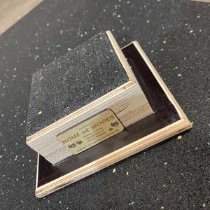 May include: A wooden ramp with a black, textured surface. The ramp is rectangular with a metal tag that reads "HOME at WOODS". The ramp is made of layered wood and has a dark border. The ramp is likely for pets.