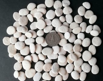 May include: A collection of smooth, white river rocks of various sizes, scattered on a dark surface. A quarter coin is placed among the stones for size comparison. The rocks have a natural, rounded shape and a slightly textured surface.