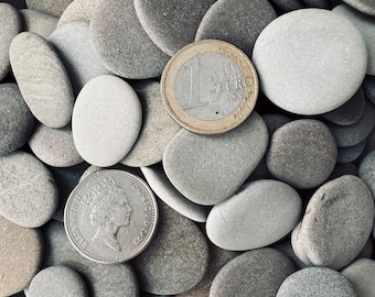 May include: A collection of smooth, gray river rocks of various sizes, with a one Euro coin and a British coin. The coins are placed among the stones, providing a sense of scale. The rocks have a natural, weathered appearance.