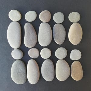 May include: Arrangement of smooth, gray river stones on a dark background. The stones vary in size and shape, with some oval and others round. The composition creates a balanced, natural aesthetic, suitable for home decor or craft projects.