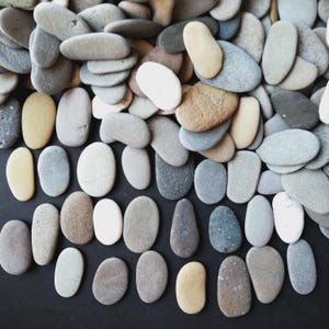 Pode incluir: Uma coleção de pedras lisas e ovais em várias cores, incluindo branco, bege, cinza e castanho claro. As pedras estão dispostas sobre uma superfície escura, algumas espalhadas e outras cuidadosamente alinhadas. As pedras naturais variam em tamanho e textura.