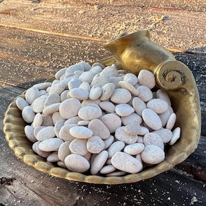 May include: A collection of smooth, matte white river stones piled inside a decorative, gold-toned metal bowl shaped like a large seashell. The bowl features a curved, horn-like handle on one side and a scalloped edge along the rim of the shell.