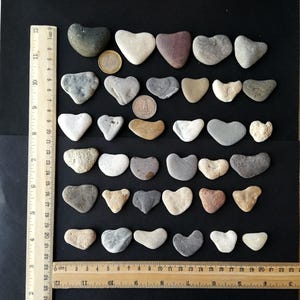 Puede incluir: Colección de piedras en forma de corazón en varios colores, grises, blancos y beiges, dispuestas sobre una superficie negra. Se observa una regla con medidas en centímetros. También hay una moneda.