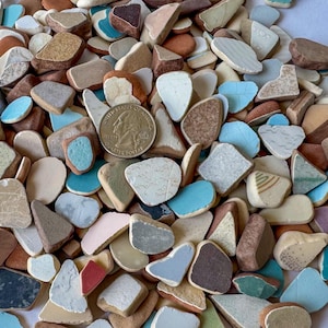 May include: A collection of colorful, irregularly shaped sea glass pieces in shades of blue, brown, tan, and white. A U.S. quarter coin is included for size reference. The pieces have various textures and patterns, creating a mosaic-like appearance.