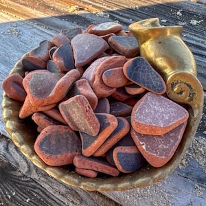 May include: A gold-toned decorative bowl with a shell-like silhouette filled with a collection of smooth, weathered sea pottery shards. The pieces are primarily terracotta red with dark blue or black glazed surfaces, showing varied shapes.