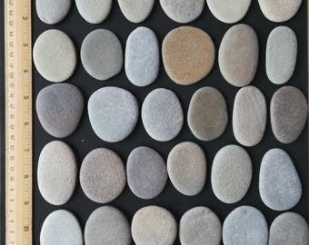 May include: An assortment of smooth, oval-shaped stones in varying shades of gray, beige, and brown, arranged on a black surface. A ruler is placed vertically along the left side, providing a size reference. The stones are likely used for decorative purposes.
