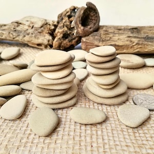 Peut inclure: Deux piles de galets beiges et lisses disposées sur une surface texturée et beige. D'autres pierres sont éparpillées autour. Des morceaux de bois flotté sont en arrière-plan, créant une esthétique naturelle et terreuse.