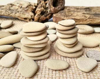 Puede incluir: Dos pilas de piedras de río lisas y beige dispuestas sobre una superficie texturizada y beige. Otras piedras están dispersas alrededor. Trozos de madera flotante en el fondo crean una estética natural y terrosa.
