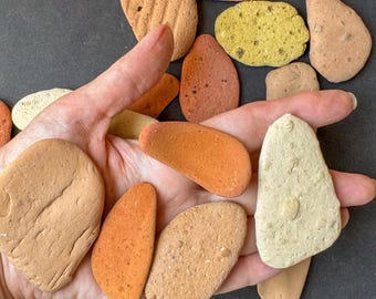 May include: A collection of various-shaped, colorful stones in shades of orange, tan, and yellow. Some stones have textured surfaces, while others are smooth. The stones are held in a hand against a dark background.