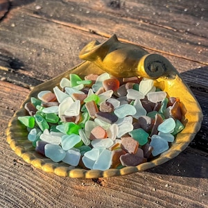 May include: A gold-toned metal dish shaped like a seashell, filled with a collection of small, smooth, frosted sea glass pieces in various shades of green, brown, white, and light blue, featuring a prominent spiral detail on the shell handle.